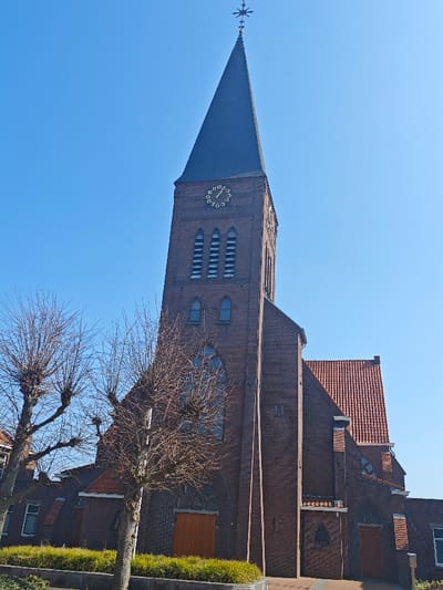An exterior, low-angle photograph depicts a red-brick church in Wierden, Netherlands, under a clear blue sky. The church features a prominent, tall steeple with a dark-grey conical roof culminating in a decorative finial. On the brick tower, a circular clock face with black numerals on a gold background is visible, indicating approximately 12:15. Below the clock, three sets of vertically oriented, louvered rectangular openings are observed, with two smaller arched windows positioned lower on the tower. The main body of the church, also constructed from red brick, has a pitched roof covered with terracotta tiles and contains large arched windows with visible tracery. Two wooden double doors are situated at ground level on the visible facade. In the foreground, two leafless deciduous trees with pollarded branches partially obstruct the lower section of the church. A low-lying green hedge is maintained along the immediate base of the building. No individuals or discernible activities are present in the scene.