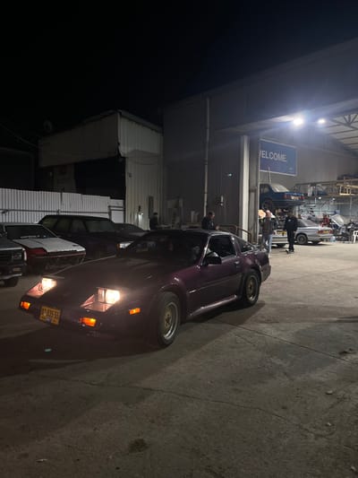 The image captures an outdoor night scene in Kfar Qasim, Israel, likely at a garage, repair shop, or industrial area. The foreground is dominated by a dark purple or maroon car, possibly a modified classic or sports car, with its headlights illuminated. This vehicle features distinctive multi-spoke, gold-colored wheels and a visible license plate displaying "91-535-55". Another vehicle, possibly a pickup truck, is partially visible behind the purple car.

The setting includes a large, brightly lit canopy structure under which various activities are taking place. In the midground, several individuals are gathered around vehicles, suggesting a car-related event, maintenance, or social gathering. A blue car is prominently elevated on a mechanical lift, indicating a repair or inspection. Other vehicles are also visible in the background.

Artificial overhead lights intensely illuminate the scene, creating strong contrasts and shadows. A blue sign under the canopy reads "WELCOME." The overall atmosphere suggests a busy, late-night session focused on automobiles. The time of day is clearly night, with the darkness of the sky contrasting with the brightly lit work area. No specific weather conditions are discernible, but the scene appears calm.