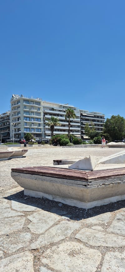 The image depicts a sunny day in Paleo Faliro, Greece. In the foreground, a concrete bench with wooden slats is prominently featured, situated on a paved area with irregular stone slabs and gravel. The bench is weathered, with visible cracks in the concrete. In the middle ground, two people, a woman and a man, are walking on the paved area. The woman is wearing a pink top and white shorts, while the man is in a light-colored shirt and shorts. Behind them, a line of palm trees and other vegetation separates the paved area from a multi-story white building with numerous balconies and windows. Further in the background, other buildings and trees are visible. The sky above is a clear, bright blue. The scene suggests a public outdoor space, possibly a promenade or a park area along the coast.