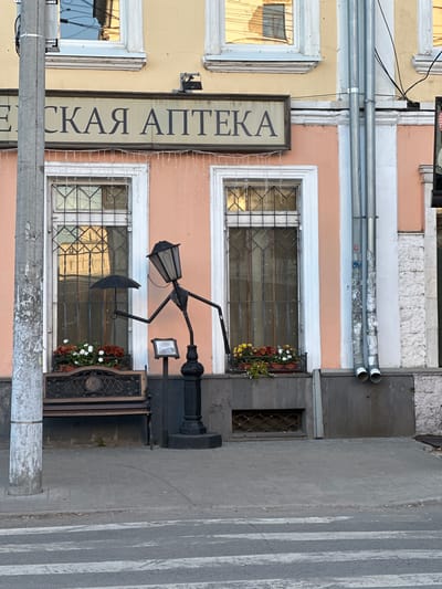 This is a photograph of a storefront on a street in Yazvitsevo, Russia. The building has a pale yellow facade on the left and a pale pink facade on the right. There are several windows on the building, some with decorative metal grates. A sign with Cyrillic text hangs vertically on the left side of the pink facade. The text appears to read "СКАЯ АПТЕКА".
In front of the storefront, there is a black street lamp with a whimsical design, resembling a figure holding an umbrella. Below the street lamp is a decorative metal grate with a circular emblem. Flower boxes with red and white flowers are visible near the windows and the street lamp. A pedestrian crossing is visible on the grey pavement to the right of the storefront. Two metal pipes run horizontally across the upper portion of the building. The overall impression is that of an old-fashioned pharmacy or shop in a European town. The lighting suggests it is daytime.