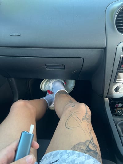 The image shows the interior of a vehicle, possibly a car, with a person's legs and lower torso visible in the passenger seat area. The person is wearing white sneakers and white socks, with Nike-branded shorts. The person has tattoos on their right knee and thigh. The person is holding a blue vaping device. The vehicle's dashboard and glove compartment are visible. There is no indication of the location of the image, and therefore the location within the city of Bristol, United Kingdom cannot be determined.
