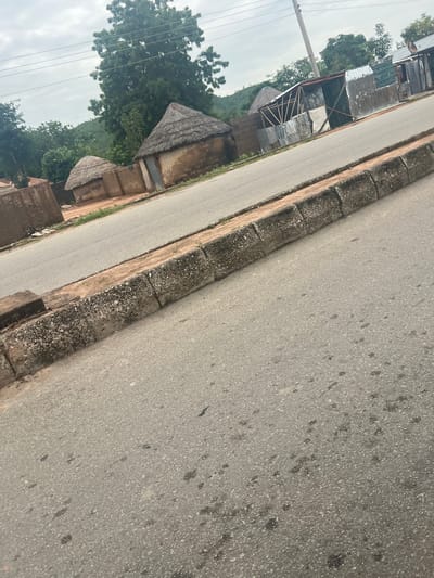 The image depicts a street scene in Kona Garu, Nigeria, likely during the daytime under an overcast sky, suggesting a cloudy or possibly rainy day. The perspective is from ground level, looking diagonally across a paved road and the adjacent sidewalk. The road surface is asphalt, showing signs of wear and tear with scattered dark stains, possibly oil or tar.
Along the left side of the road, a row of structures lines the street. These buildings appear to be a mix of traditional and more modern constructions. Some have thatched roofs, characteristic of older dwellings, while others are covered with corrugated metal sheets, suggesting recent additions or repairs. One building, partially visible, has a wooden frame supporting a metal roof. In front of these buildings, there's a clear demarcation between the road and the properties, formed by a rough concrete curb or low wall. The area behind the buildings seems to be a vegetated hillside.
The overall atmosphere is that of a rural or semi-urban settlement. There are no people visible in the image, and the focus is on the built environment and the natural surroundings. The presence of utility poles and overhead wires suggests that electricity is available in this location. The lush green trees in the background indicate a tropical or subtropical climate. The image conveys a sense of everyday life in a Nigerian village or town, with a blend of traditional and contemporary building materials.
