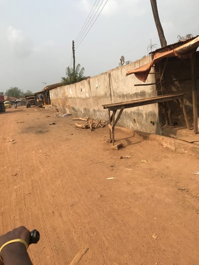The image depicts an unpaved reddish-brown dirt road in Ogbese, Nigeria, under a hazy light-colored sky. The perspective is from the immediate foreground, with a partial view of a dark-skinned arm wearing a yellow wristband and holding a black handlebar-like object in the bottom-left corner.

Along the right side of the road, a long, weathered concrete wall extends into the mid-ground. Razor wire or barbed wire is affixed to the top of the wall in sections. Adjacent to the wall, a makeshift wooden stall with a corrugated metal roof covered by an orange tarp is visible. In front of this, a simple wooden table structure stands on the dirt. A pile of cut logs or firewood is located on the ground between these structures and the road. Scattered debris and litter are present on the ground.

On the left side of the road, utility poles with overhead power lines are visible. Further down the road, several structures with corrugated metal roofs line the street. Vehicles, including a yellow three-wheeled auto-rickshaw (Keke Napep) and a large dark truck, are parked or in motion. Multiple indistinct figures are visible near the vehicles and buildings in the mid-ground. Sparse green vegetation is visible behind the wall and structures in the distance.