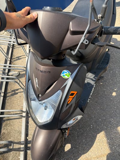 A person's hand, tattooed on the back with a faded design, grips the handlebars of a brown Kymco scooter parked in front of a metal bike rack. The scooter is positioned on an asphalt surface, with gravel and sand visible. The environment appears to be outdoors, possibly a public area, given the bike rack. The scooter features prominent "KYMCO" branding, a circular decal with green and blue elements, and an orange graphic with the number "50". The sunlight casts distinct shadows, indicating a bright, clear day. The image is taken from a slightly elevated, forward-facing perspective, focusing on the front of the scooter and the person's hand.