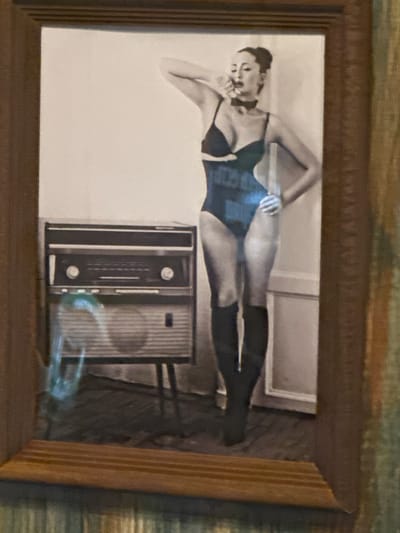 The image shows a framed black and white photograph. The photograph depicts a woman reclining in a one-piece bathing suit and knee-high boots next to an old radio. The location of this image is in Selyatino, Russia.