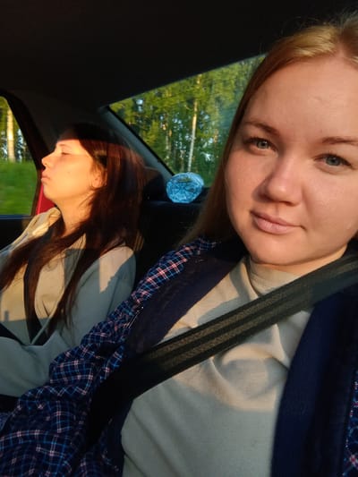 Two individuals are visible inside a vehicle traveling through a rural area, possibly on the outskirts of Bogorodsk, Russia. The person in the foreground is a young woman with blonde hair, looking towards the camera. She is wearing a light-colored t-shirt and a dark blue jacket with a plaid pattern. A seatbelt is fastened across her chest. The second individual, in the background and to the left, is a young woman with dark brown hair, appearing to be asleep with her eyes closed. She is also wearing a light-colored hoodie and a seatbelt. The interior of the car is dimly lit, with sunlight streaming in from the side, casting shadows and illuminating the scene. Outside the car windows, trees and greenery are visible, suggesting a natural, possibly wooded environment. A partially obscured blue object, possibly a bottle, is visible on the back seat between the two individuals. The overall impression is a candid snapshot of a road trip, capturing a moment of rest and travel during daylight hours.