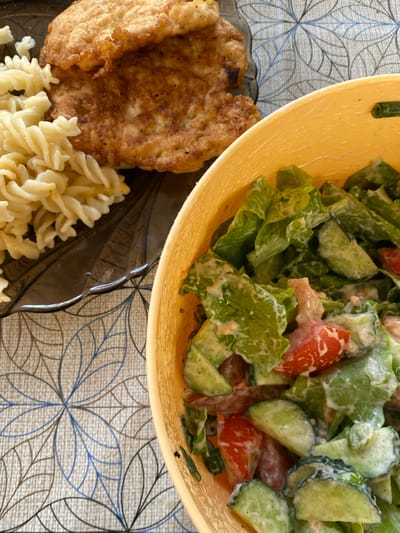 The image shows a close-up, overhead view of a meal on a patterned tablecloth. On the left, a dark plate holds a piece of fried chicken or schnitzel next to a serving of spiral pasta. To the right, a yellow bowl contains a salad made with lettuce, sliced cucumbers, and chopped tomatoes, all coated in a creamy dressing. The tablecloth has a repeating pattern of stylized blue and brown floral motifs. The scene appears to be indoors, possibly in a dining area. There are no people or specific landmarks visible that would indicate the location in Velykyi Bychkiv, Ukraine.