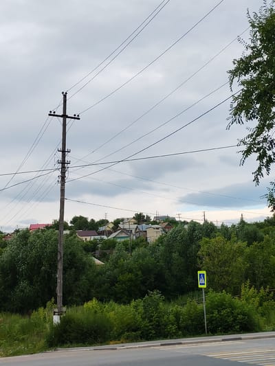 This media file depicts a somewhat overcast, daytime scene in Volsk, Russia, likely in a residential area on the outskirts of town. The focal point is a wooden utility pole with multiple electrical wires extending from it, crisscrossing the frame against a cloudy sky. Behind the pole, a densely wooded hillside is visible, dotted with various houses, suggesting a village or suburban setting. Some houses have brightly colored roofs, adding a touch of contrast to the predominantly green foliage and gray sky.
In the foreground, the edge of a paved road is visible, with a pedestrian crossing marked by yellow and white stripes. Beside the road, a small, square blue and yellow sign with a pedestrian symbol indicates a crosswalk. The lush green trees and undergrowth dominate the lower half of the image, indicating a temperate climate and likely summer or late spring. There is no visible activity or people in the scene, giving it a quiet, almost still atmosphere. The lighting suggests it is daytime, but the clouds create a diffused, muted light, preventing strong shadows. The overall impression is a functional, somewhat rustic view of a Russian provincial town, blending infrastructure with natural elements.