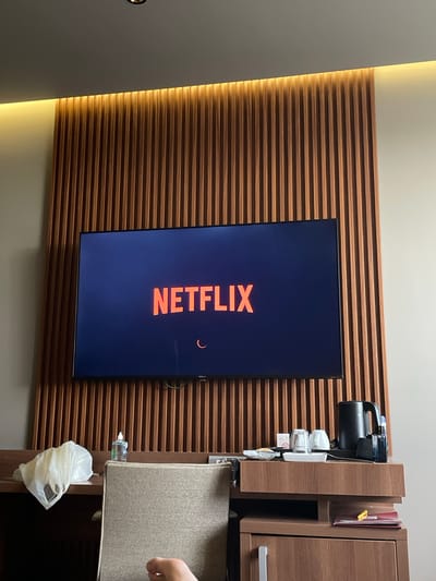 A residential interior in Otopeni, Romania, features a wall with horizontal wooden slats. Centered on the wall is a large, vertically-oriented television screen displaying the Netflix loading screen. To the right of the television, a dark brown cabinet with a side compartment holding items, including a black coffee maker and various cups. Below the cabinet, an extending, fabric-covered surface is partially pulled out, with a white plastic bag hanging from a clear bottle attached to its side. A partial view of a hand is visible on the right edge of the extending surface. A portion of a white wall is seen to the left of the slatted wall. The lighting in the room suggests both natural and artificial sources, highlighting the wooden slats.