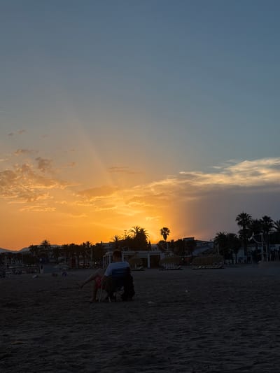 A photograph taken at sunset on a beach in Cambrils, Spain. The image is oriented vertically. The sky transitions from a pale blue on the left to a vibrant orange and yellow sunset on the right, with scattered clouds. Silhouetted palm trees and buildings line the background, suggesting a resort or coastal town. The foreground is a sandy beach with ripples from the wind or tide.

In the middle ground, a person is sitting on a folding chair, facing away from the camera, with a backpack beside them. The person appears to be wearing a white shirt and dark shorts. The setting sun casts long shadows across the sand. The overall mood is peaceful and serene, characteristic of a beach at dusk. No visible text is present in the image. The lighting indicates it is late evening.