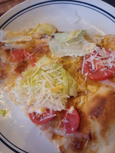 A close-up, overhead shot shows a slice of pizza with lettuce, tomatoes, and shredded cheese on a white plate with blue stripes. The pizza appears to be a cold slice as the lettuce is fresh and not wilted from heat. The lighting is bright and even, casting no discernible shadows. The image is focused on the food item, with the background being a blurred wooden surface. There is no indication of location in Ukrainka, Ukraine or any people present.