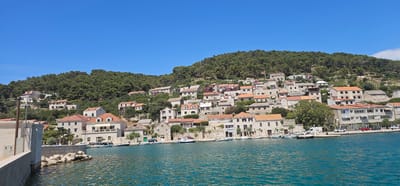 A wide, panoramic view captures a coastal village nestled on a hillside under a clear blue sky. The village, identified as being in Općina Pučišća, Croatia, features a dense cluster of buildings with terracotta tile roofs cascading down towards the azure water. The architecture is characteristic of the Mediterranean, with many structures made of stone and painted in light, neutral colors. Some buildings have distinctive green shutters, adding pops of color to the scene.
In the foreground, the sparkling blue water of the sea laps gently. A concrete seawall or pier, partially visible on the left, extends into the frame. Several small boats are moored along the waterfront, some of which are dinghies and others larger leisure craft. Behind the boats, a paved promenade or street runs parallel to the coastline, with parked cars visible.
The hillside rising behind the village is densely covered with lush green trees, predominantly pines, creating a stark contrast with the built environment. The overall impression is one of a tranquil, sun-drenched coastal settlement. There are no people prominently visible in the image, contributing to the peaceful atmosphere. The scene is bathed in bright sunlight, suggesting it is a clear, warm day.
