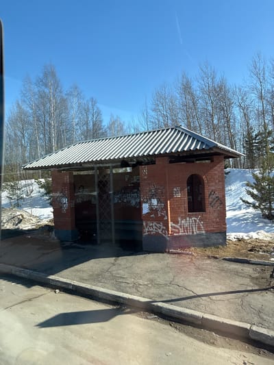 The image depicts a red brick bus stop shelter with a corrugated metal roof, situated on the side of an asphalt road in Bolgury, Russia. The structure features a dark gray or black painted base, and its red brick walls are extensively covered with white and black graffiti tags and torn paper posters. One side of the shelter shows an arched, window-like opening. Inside the shelter, a simple frame or support structure is visible, and debris is present on the ground.

Surrounding the bus stop, patches of melting snow and exposed dry earth/vegetation are visible, suggesting a late winter or early spring season. Behind the shelter, a snow-covered embankment rises, populated by numerous bare deciduous trees, possibly birches, with a few small evergreen conifers also present. The sky above is clear and bright blue, indicating sunny weather. A distinct shadow from the bus stop structure falls across the cracked asphalt surface in the foreground, where a concrete curb separates the road from the bus stop area. No individuals or active interactions are visible within the scene.
