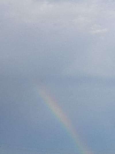 A faint rainbow arc is visible against a hazy, overcast sky. The rainbow appears in the lower left quadrant of the image, extending upwards and to the right. The sky is a muted blue-gray, with some lighter cloud formations in the upper portion of the frame. There are faint horizontal lines visible near the bottom of the image, possibly power lines, suggesting a landscape or urban environment. No people or specific objects are discernible within the rainbow or the sky. The overall impression is of a soft, diffused light. The location is stated as Yar-Bazar, Russia.