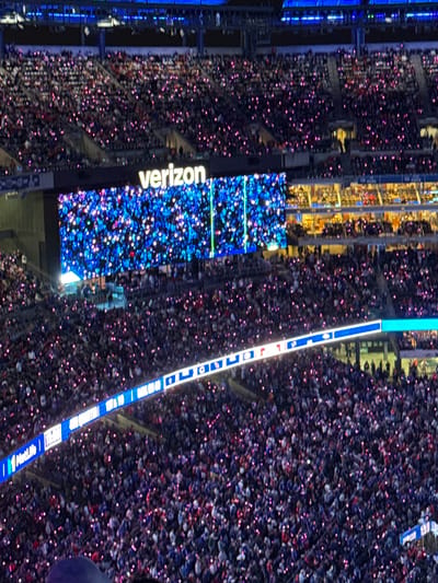 This image depicts the interior of MetLife Stadium, located in East Rutherford, New Jersey, United States, during a nighttime event. Thousands of spectators are visible occupying densely packed, tiered seating sections. A large majority of these individuals are holding up devices that emit small, distinct pinkish-purple lights, creating a scattered pattern of illumination across the entire visible audience.

In the mid-ground, a prominent, large rectangular video screen displays the word "verizon" in white text above an animated graphic consisting of numerous blue and white "sparkle" effects on a dark background, with two vertical green lines on its right side. Below this main screen and spanning across a lower seating tier, a horizontal LED ribbon board with a blue border exhibits various illuminated graphics and text, including a "MetLife" logo visible on its far left.

The overall stadium environment is dimly lit, with ambient blue lighting illuminating upper structural elements and some warmer, yellowish light emanating from enclosed box seats situated behind and to the right of the main video screen. The scene conveys a large-scale public gathering characterized by collective audience participation in a light display.