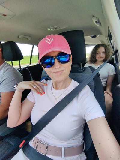 The image depicts three individuals in the interior of a car, likely traveling through the rural landscape surrounding Дзяржынскі сельскі Савет, Belarus. A woman in the foreground, wearing a pink baseball cap with a white heart and blue mirrored sunglasses, is taking a selfie. She has bright orange nail polish and a light pink t-shirt. A silver necklace with a pendant is visible. She is wearing a watch on her left wrist. A second person, a young female with curly brown hair, is seated in the back passenger seat, wearing a white t-shirt. Her seatbelt is visible. A third individual, partially visible on the left side of the frame, appears to be a male wearing a grey t-shirt. The interior of the car includes car seats, headrests, and seatbelts. The windows show a view of a green, hilly landscape, consistent with the rural surroundings of Дзяржынскі сельскі Савет. The overall lighting suggests it is daytime.