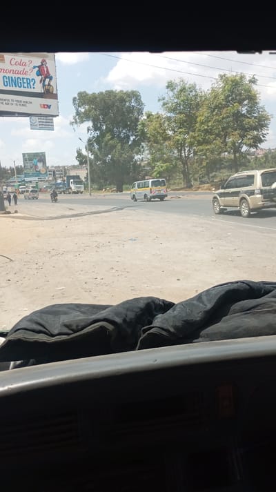The image captures a road scene from the perspective of someone inside a vehicle, looking out through the front windshield on a bright, sunny day. The bottom portion of the frame shows the dark dashboard of the vehicle and a black, rumpled jacket or fabric resting on it.
In the midground, a dusty, unpaved shoulder on the left transitions into an asphalt road. On the asphalt, several vehicles are visible. A gold or beige SUV, possibly a Mitsubishi Pajero, with a spare tire mounted on the rear, is driving away from the camera on the right side of the road. On the left side of the road, a white and yellow minivan, resembling a public transport vehicle (common in some African countries), is also driving away. Further down the road on the left, another white van and a large white truck are parked or moving slowly. A person on a motorcycle is visible near the white van.
On the unpaved shoulder to the far left, two individuals are walking away from the camera. In the mid-distance, beyond the road and near some trees, several animals that appear to be goats or sheep are grazing or standing.
The background features numerous green trees, suggesting lush vegetation. Beyond the trees, some distant buildings and structures are visible, indicating a semi-urban or rural setting. The sky is partly cloudy with significant blue patches, indicating clear weather.
On the far left, two billboards are prominently displayed. The upper billboard shows a cartoon-like drawing of a person on a unicycle. Text on this billboard reads: "Cola? Lemonade? GINGER? HARMFUL TO YOUR HEALTH. HE AGE OF 18 YEARS UDV". Below this, there are three phone numbers: "074655282", "0754230051", and "0771830144". The lower billboard is partially obscured but shows an image of people and some text, likely advertising a product or service.