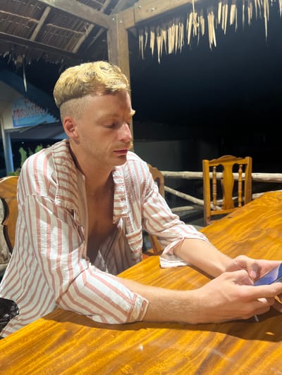 An overhead shot shows a man with blonde, wavy hair and a mustache sitting at a wooden table. He is wearing a pink and white striped button-down shirt, unbuttoned at the top, revealing his chest. The man is looking down at a smartphone in his hands. The background is dark, but a thatched roof and a wooden chair are visible. The image appears to have been taken at night at a restaurant in Thung Tako, Thailand, possibly on a beach due to the thatched roof structure.
