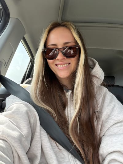 A young woman with long brown hair and blonde highlights, wearing sunglasses and a light grey hoodie, is in a car and wearing a seatbelt. The car appears to be in motion, and sunlight is visible through the window. The location is unknown and can't be determined from the image. Ternovka, Russia cannot be determined from the image.