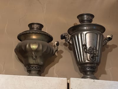 The image displays two antique samovars, traditional Russian metal containers used for heating and boiling water for tea. They are positioned on a white surface against a textured, light brown wall. The samovar on the left is made of a brass-like metal and features ornate decorative elements, including a fluted body and a decorative band around its base. The samovar on the right is made of a darker, silver-toned metal with vertical fluting and a spigot for dispensing water. Both samovars have handles and decorative lids. The lighting suggests an indoor setting, possibly a home or museum, with the light casting shadows on the wall behind the objects. There is no visible activity or event taking place, and no people are present. The overall impression is one of historical artifacts displayed in a static setting. Some of the decorative bands on the samovars appear to have a repeating pattern that could be interpreted as text or symbols, but it is not clearly legible. The location context provided is "Патрушева, Russia."