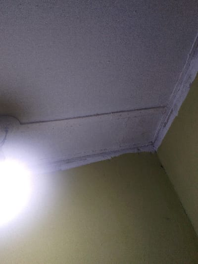 A dim interior corner shows a textured ceiling with a rectangular border detail. The walls, painted in a faded olive green, meet at a sharp angle. The ceiling is likely white or off-white. A bright, out-of-focus light source obscures the lower left corner, casting a glare across the scene. The overall image has a slightly grainy texture and a somewhat uneven lighting distribution.