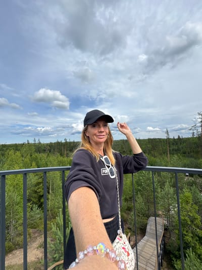 A woman with blonde hair, wearing a black baseball cap, black sweatshirt, and black shorts, stands on an elevated platform overlooking a forest. She is holding her phone out to take a selfie. She has white sunglasses hanging from the neck of her sweatshirt and a patterned purse slung over her shoulder. A beaded bracelet is visible on her left wrist. The background shows a dense green forest with scattered tall trees under a cloudy sky. To the right of the woman, a wooden walkway with railings descends into the forest. The location is the Peski nature reserve in Russia.