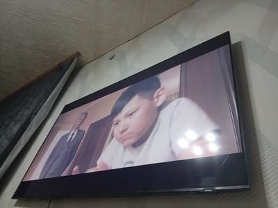 A wide-angle, slightly tilted, medium shot captures a modern, flat-screen television displaying a scene. On the left side of the television screen, a man in a dark suit and tie stands, looking towards the right. To the right of him, a young boy with dark hair and wearing a light-colored shirt is visible, his face close to the camera and appearing to be speaking or reacting to something. The background on the television screen shows an interior setting with wooden doors. The television itself is dark with a thin black bezel, and a "SAMSUNG" logo is visible on the bottom bezel. The television is mounted on a wall covered in patterned wallpaper with a textured, embossed design. To the left of the television, a dark, pleated fabric hangs down. The scene is illuminated by ambient light, creating reflections on the television screen, and shadows are cast on the wall. The image is taken from a low angle looking upwards. The location is identified as Kastornoye, Russia.