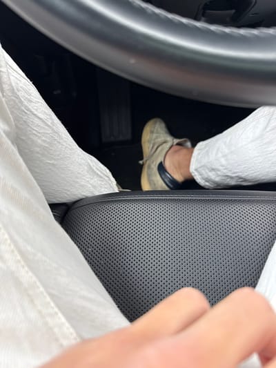 The image forensically details the interior of a vehicle, capturing a scene from the driver's perspective in Wellesley, United States. The lower body of an individual is visible, wearing white, textured pants. The subject's right foot, clad in a light-colored, possibly beige or grey, lace-up sneaker with a dark blue or black heel accent, is positioned above the accelerator pedal. A dark, perforated leather car seat is prominently featured in the lower-mid foreground. The bottom segment of a dark steering wheel, characterized by a metallic-toned rim, is visible in the upper portion of the frame. A hand is partially visible at the bottom center of the image. The vehicle's dark floorboard and additional pedals are discernible in the footwell.