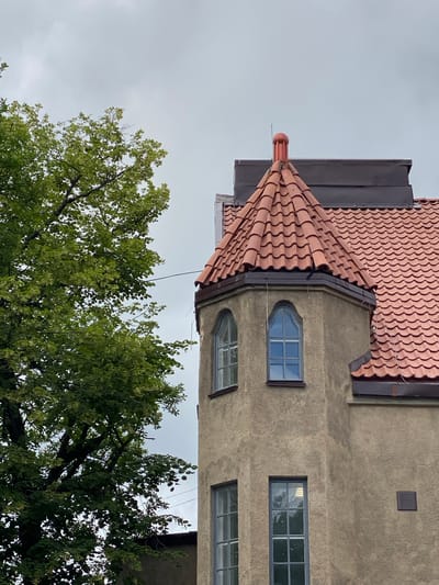 The image shows a detail of a tan stucco building with a red tiled roof, featuring a turret with arched windows. A large green tree with dense foliage is positioned to the left of the building, partially obscuring the view of the structure. The sky is overcast with a light gray cloud cover. The scene is likely captured on a day with diffused light, typical of a cloudy or rainy climate. The building's architecture suggests it could be an older structure, possibly in a historical district or a residential area. The presence of the tree and the building's texture create a sense of depth and architectural interest.