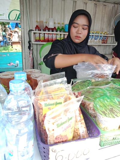 The scene depicts a woman wearing a black hijab at a food stall. She is arranging a clear plastic bag, likely containing food items, on the stall. Displayed are various packaged goods, including "Makaroni Goreng Lywa" and containers with green leaf-patterned lids. Bottles of water and larger containers of tea are visible in the foreground. In the background, shelves hold bottles of syrups and packaged drinks. The stall has a metal structure, and outside, a glimpse of a street scene with a blue motorcycle is visible. The price of an item, "6.000", is written on the stall. The location is likely Kebondalem, Indonesia.