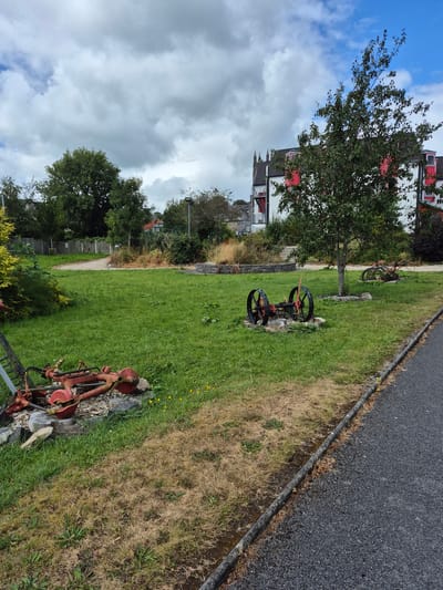 The image depicts an outdoor scene with a grassy area, a paved path, and a building in the background. In the foreground, to the left, lies a dismantled piece of red agricultural machinery on a bed of rocks and wood. Further into the grass, a complete piece of similar red machinery with two large spoked wheels and a handle is displayed. In the background, a multi-story building with white walls and red trim is visible, featuring dark roofs and a prominent red fire escape. The building appears to be set in a park-like environment with trees and shrubs. The sky is cloudy with patches of blue. The scene is located in Carrick-on-Shannon, Ireland.