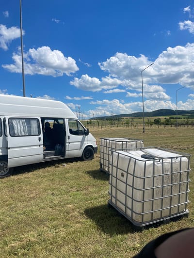In a grassy field with scattered trees and hills in the background, two large white plastic water tanks with metal cages are present. To the left, a white van with its side door open is parked. The van has curtains in its windows and appears to be empty inside, except for some bottles. The sky is blue with scattered white clouds. Streetlights and power lines are visible in the distance. The location is Neofit Rilski, Bulgaria.
