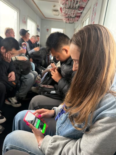 The image captures a candid, slightly tilted view inside what appears to be a public transport vehicle, such as a bus, train, or ferry, in Nizhneleninskoe, Russia. The interior features white walls, windows, and rows of seats, some with red cushioning visible on the left.

The foreground is dominated by a person with long, wavy brown hair, wearing a denim jacket over a grey hoodie. This individual is engrossed in a bright red-cased smartphone, held with both hands. The phone screen displays a vibrant green interface with various numbers and what appears to be English text, possibly related to a game, social media, or a tracking application. The person's fingernails are painted bright pink.

To the left of the foreground subject, another person with short, dark hair is visible, also looking down, likely at a phone. The scene is populated by numerous other individuals, mostly young adults or teenagers, seated in rows, stretching into the mid-ground. Many are looking down, suggesting they are occupied with their personal devices, while others have their heads resting or are looking outwards. Their faces are largely obscured or blurred.

Clothing details include various jackets (denim, dark puffer jackets), hoodies, jeans, and sneakers. Backpacks and bags are also visible among the seated passengers. Light streams in through the windows, indicating it is daytime. The overall atmosphere suggests a quiet, focused engagement with personal devices during travel. Visible text on the phone screen is difficult to fully decipher but includes numbers and English words on a green background. Blurry signs or symbols are also present on the wall above the windows in the background.