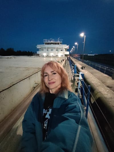 The photograph captures a selfie taken by a woman with shoulder-length auburn hair and fair skin, wearing a teal-colored jacket over a black t-shirt. She is positioned in the foreground, looking directly at the camera with a slight smile. The background features a stark, concrete pathway illuminated by a series of streetlights. Beyond the pathway, a large, multi-story white building with numerous windows is visible, appearing to be a structure related to water transport, possibly a lock or terminal. The sky above is a deep twilight blue, indicating the image was taken during dusk or early night. The overall scene is set outdoors, likely in an industrial or port area. The location is identified as Sheksna, Russia, suggesting a connection to the Mariinsk Canal system or a related waterway.