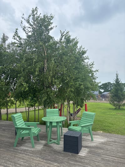 An outdoor seating area is depicted on a wooden deck in a cloudy, overcast setting, possibly in Tavrichanka, Russia. The scene features a round green table and four green chairs, resembling Adirondack style, arranged in a conversational grouping. A dark grey cube-shaped object, possibly a stool or small table, sits near one of the chairs. Behind the seating area, a line of slender birch trees with lush green foliage dominates the mid-ground, their trunks appearing light-colored. The background transitions to a manicured green lawn dotted with more trees, including a conical evergreen on the right. Faint structures, possibly buildings or sheds, are visible in the distance. A red vertical post, likely part of an unseen structure or play equipment, is visible between the trees. A hammock appears to be strung between two unseen supports behind the trees. The wooden deck shows signs of moisture, suggesting recent rain. No people are present in the image.