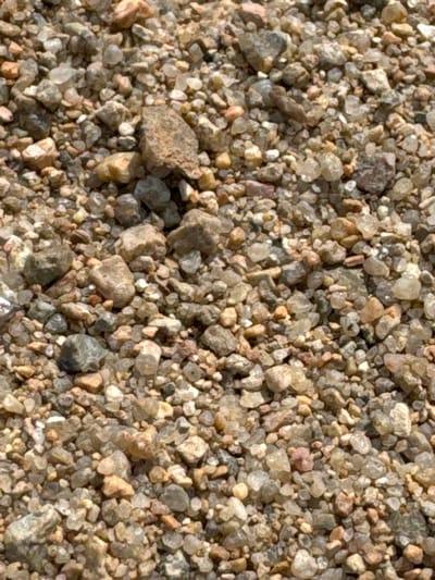 The image shows a close-up, overhead view of a pile of small pebbles and sand. The pebbles are a mix of colors including grey, brown, white, and some reddish-pink hues. They vary in size from fine grains of sand to pieces roughly 1-2 centimeters in diameter. The texture is rough and granular. The lighting suggests an outdoor environment. There are no people or discernible structures visible. The image is a generic depiction of loose aggregate material and does not contain specific details that would allow for identification of its location within Ayyrtau, Kazakhstan.