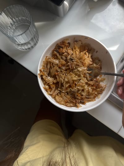 A top-down view shows a white bowl filled with cooked cabbage and carrots, being stirred by a fork held by a person's hand. To the left of the bowl, a clear glass with water sits on a white surface, possibly a kitchen counter. The background is dark, with a hint of a metallic appliance. The scene is lit by natural light coming from the right, casting shadows. There is no indication of the location being Baan Nok Sai, Thailand.