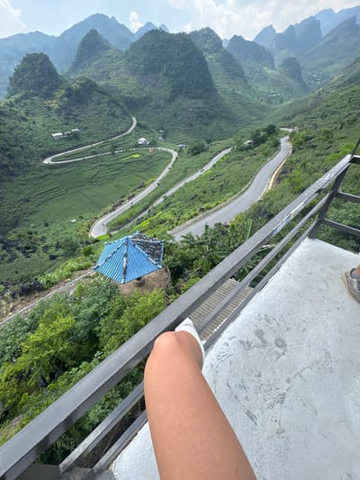 An elevated shot overlooks a sinuous mountain road near Lũng Phìn Commune, Vietnam. The shot is taken from a high viewpoint, likely a structure with a gray metal railing and a concrete base, visible in the frame. A leg wearing a white sock rests against the railing. Below, the landscape is characterized by verdant hills, terraced fields, and scattered buildings. A structure with a prominent blue tiled roof stands out. The road winds through the landscape, ascending and descending the hills. In the distance, layers of mountains are visible under a sky with scattered clouds.