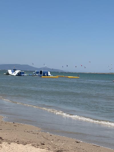 The image captures a sunny day at a beach with clear blue skies and calm, shimmering water. The scene is filled with various kitesurfers in the distance, dotting the horizon and adding a splash of color to the vast expanse of the sea and sky. In the foreground, several inflatable water structures are partially submerged near the shore. One prominent structure is a large, blue and white inflatable platform with a pipe-like feature and what appears to be a number "7" visible on its side. Another smaller, white inflatable is also present. The gentle waves are lapping at the sandy shore, where small shells and debris are scattered. The overall impression is one of a leisurely summer day at a coastal location, likely Greece given the context. The sunlight glinting off the water suggests it is midday.