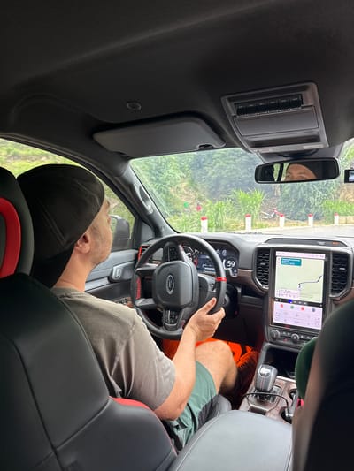 The image captures the interior of a modern Ford Raptor vehicle, viewed from a rear passenger seat looking forward. The photo is rotated 90 degrees clockwise. A male driver, wearing a dark cap, grey t-shirt, green shorts, and orange shoes, is seated in the driver's seat, holding the steering wheel with his right hand.
The dashboard features a prominent, vertically oriented central infotainment screen displaying Google Maps navigation, showing a route with an estimated time of 13:02 and a temperature of 22.5°C. Climate controls are visible below the screen. The digital instrument cluster behind the steering wheel shows "59," likely a speed reading. The steering wheel is branded with "FORD" and "RAPTOR."
An overhead console contains six auxiliary switches labeled "AUX1" through "AUX6." The rearview mirror reflects a person, possibly female, wearing a dark head covering, suggesting the photographer. Through the windshield, green foliage and several red and white striped poles are visible, indicating an outdoor setting, potentially a path or trail in Minh Sơn Commune, Vietnam. The gear shifter is a rotary dial. The vehicle's interior is dark-toned with black seats featuring red accents. The scene takes place during bright daylight.
