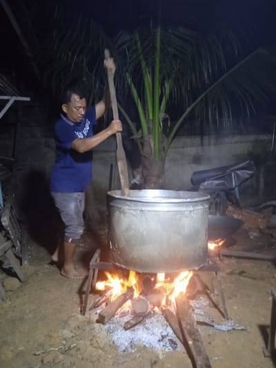 A middle-aged man is stirring a large metal pot with a wooden ladle over an open fire in Keudeteupinpunti, Indonesia. The man is wearing a blue t-shirt and grey pants. The pot is supported by a metal stand, and firewood is burning beneath it. A palm tree is visible in the background, and the scene is dimly lit, suggesting it is nighttime. Other objects in the background are obscured by darkness. The ground is covered with dirt and debris.