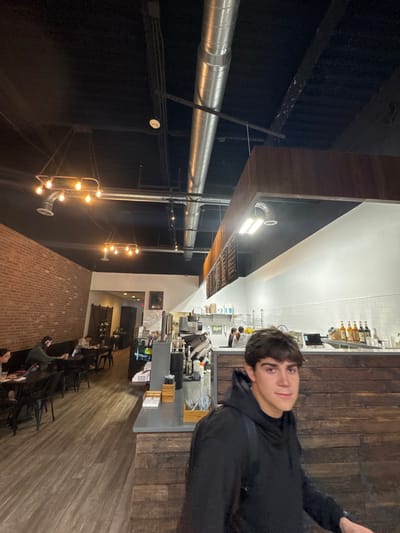 The image depicts the interior of Zen Cafe, a coffee shop located in South Bend, Indiana, United States. A young male with dark hair, wearing a black hooded sweatshirt and backpack, is positioned in the right foreground, looking towards the camera. The establishment features a black ceiling with exposed silver HVAC ductwork and electrical conduits. An exposed red brick wall runs along the left, illuminated by warm-toned, multi-bulb industrial pendant lights. The floor is light-colored wooden laminate. In the left midground, a customer seating area with dark tables and chairs is occupied by three individuals, primarily engaged with laptops. To the right, a service counter with a dark grey surface and wooden plank facade displays coffee equipment, various bottles, and supplies. Behind this counter, against a white subway tile wall, two employees are visible. Dark wooden menu boards are mounted overhead above the counter.