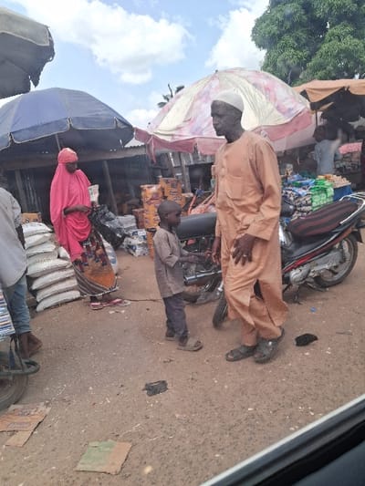 The image depicts an active outdoor market scene in the city of Kungana, Nigeria, under a partly cloudy sky. The ground is unpaved, appearing dusty and uneven, with scattered debris including pieces of cardboard.
In the foreground to mid-ground, four individuals are visible. On the right, a dark-skinned adult male stands, facing left, wearing a light orange long-sleeved garment (kaftan) and a white skullcap. He wears dark sandals and has his left hand slightly raised. To his left, a dark-skinned young male child stands, facing the man, wearing a light brown long-sleeved shirt, dark pants, and light-colored sandals. The child extends his right hand towards the man, suggesting an interaction. Between them and slightly behind, a dark-colored motorcycle or scooter is parked on its kickstand, facing right.
To the left of the child, a dark-skinned adult female is visible, facing right. She wears a bright pink head covering (hijab) and a matching pink flowing garment over a patterned skirt or wrapper. She holds a dark bag with her right hand and appears to be sorting or handling items. Immediately to her left, a stack of white sacks is visible, and the lower legs and feet of another individual, wearing blue jeans and dark flip-flops, are partially visible near a bicycle wheel.
In the background, various market stalls are present, characterized by corrugated metal structures and large shade umbrellas. A large blue umbrella is on the left, and a large patterned pink and white umbrella is centrally located behind the man and child. Further back, a smaller brown umbrella is visible. Stacks of packaged goods, including cardboard boxes and plastic-wrapped items, are visible behind the individuals and under the market structures. A large tree with green foliage is visible on the upper right, contributing to the open-air environment.