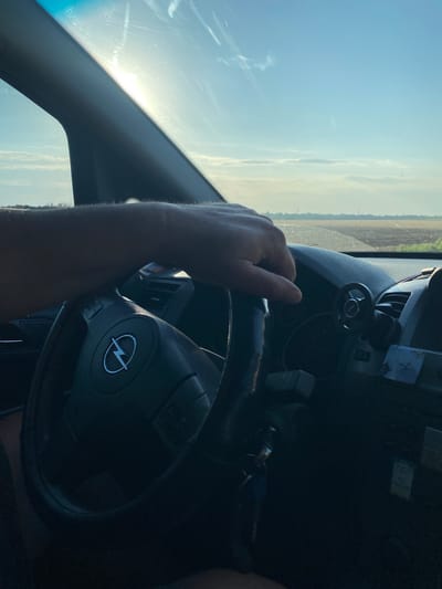 An over-the-shoulder shot captures a person's left hand resting on the steering wheel of a car, as seen from the driver's perspective. The steering wheel has the Opel logo embossed on it. Beyond the windshield, a rural landscape unfolds under a bright, sunny sky with scattered clouds. The horizon features fields and what appear to be utility poles. The scene is set on a road outside of Maiske, Ukraine. The interior of the car shows part of the dashboard and a small, circular accessory attached to it.