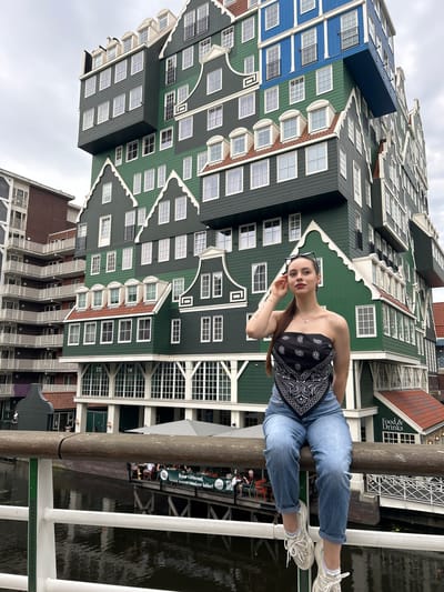 A young woman sits on a wooden railing in front of the Inntel Hotel Zaandam, Netherlands. The hotel's distinctive facade, composed of stacked green, blue, and grey segments designed to resemble traditional Zaandam houses, dominates the background. The woman is wearing a black bandana as a top, blue denim jeans, and white sneakers. She is looking slightly to her left and touching her sunglasses, which are perched on her head. Below the railing, a canal is visible, with a pedestrian walkway and outdoor seating area of a restaurant on the opposite bank. The sky is overcast.