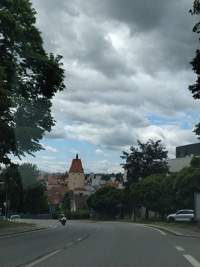 The image captures a view of a city street with a motorcycle rider in the foreground, approaching from the distance. The rider is on a motorcycle, wearing a helmet and protective gear. The street curves to the right, with a white van parked on the left side of the road. Beyond the road, a cluster of buildings with red-tiled roofs is visible, dominated by a tall, light-colored tower with a red roof and a clock face. The sky is overcast with dramatic, dark clouds. Trees line the sides of the road and frame the cityscape. The scene appears to be in a European city, with architectural styles suggesting a location in the Czech Republic, possibly Olešná given the prompt. A silver SUV is parked on the right side of the road near a dense cluster of trees.