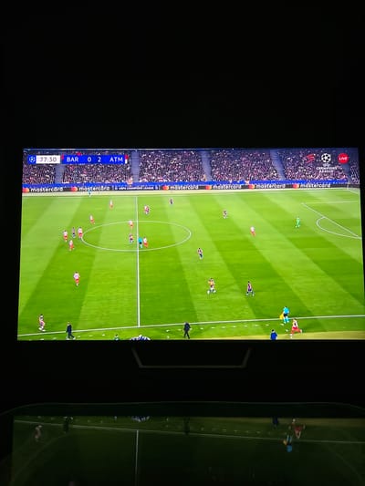The image shows a television screen displaying a live broadcast of a UEFA Champions League football match. The score is Barcelona 0, Atletico Madrid 2. The time on the clock is 77:30. The field is green with white lines marking the boundaries and center circle. Players are visible on the field, wearing different colored jerseys, in the midst of game play. Crowd is visible in the background. The broadcast includes Mastercard advertising around the stadium. There is nothing in the image to indicate the city of Germiston, South Africa.
