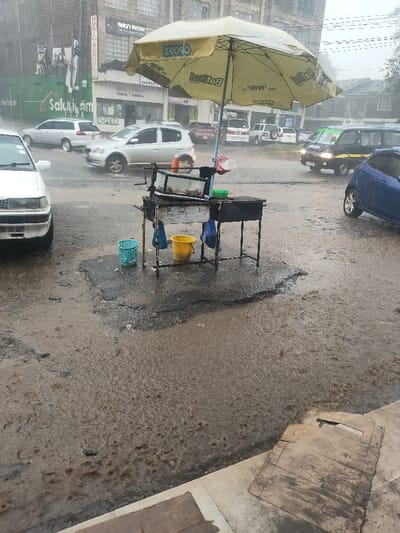The image depicts an urban street scene in Oloolua ward, Kenya, under active rainfall. A yellow promotional umbrella, branded with "29090" and "Betika," shelters a metal street vendor's stall in the mid-ground. The stall features a display unit, possibly a grill, with items visible inside a clear partition. Below the stall are a light blue and a yellow plastic bucket, with two blue plastic bags hanging from the frame. The asphalt road surface is thoroughly wet, showing numerous puddles and visible rain impact ripples. A white sedan is parked on the left foreground. In the mid-ground, a silver hatchback is in motion towards the left, displaying visible motion blur on its wheels. Further into the scene, a dark minivan and a blue hatchback are also on the wet road, with additional vehicles parked further back. Commercial buildings line the background, featuring various signs, including "Music School," "Micky's Parlour," "Safaricom," and "MPESA." No individuals are visible in the frame.