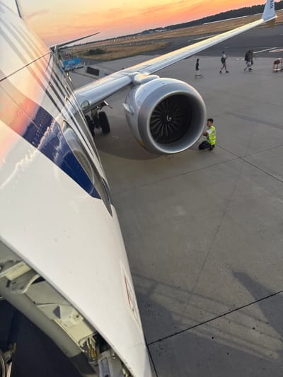 An external shot taken from the perspective of a passenger on an airplane at an airport, likely in Weeze, Germany, during sunset. The aircraft's fuselage is prominently in the foreground on the left, showing its white exterior with blue stripes. To the right, a large jet engine is visible, with its intricate fan blades exposed. A person wearing a high-visibility yellow vest is crouched near the engine, appearing to conduct an inspection or maintenance. In the background, beyond the wing and runway lights, people are walking, and the sky is painted with the warm hues of a sunset. The scene suggests an operational airport environment at the end of the day.