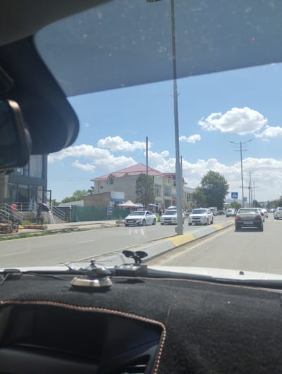 A street view from inside a vehicle looking towards a road with traffic in Forish QFY, Uzbekistan. The sky is blue with scattered white clouds. On the left side of the road, there is a building with glass windows and signage that reads "JASMIN". A person holding a purple umbrella walks on the sidewalk in front of this building. Further down the street, there are more buildings, trees, and power lines. Several cars are driving on the road, including white sedans and a silver car. The foreground shows the dashboard of the vehicle, with a small silver helicopter ornament. The road has a yellow and grey curb.